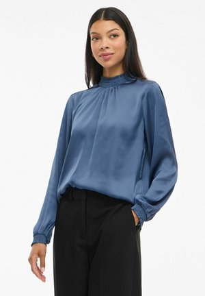 Vrouw met lang, donker haar draagt een blauwe satijnen blouse met een volants hoge hals en manchet, in combinatie met zwarte broek, staand met één hand in de zak.