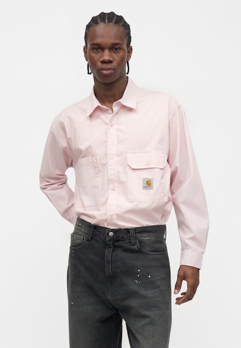 Young man with braided hair wearing a light pink long-sleeve button-up shirt and black jeans with white paint splatters.