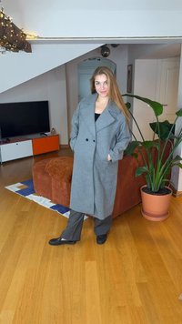 Grå ullkappa med dubbelknäppning, vida slag och sidfickor, bärs över mörka byxor, framför en modern inredningsbakgrund.