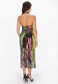 Nainen, jolla on yllään monivärinen paljettimidi mekko ja mustat avokkaat nilkkaremmeillä, sekä rannekoru, seisoo selin.