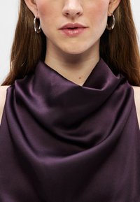 Fialový saténový top s překrytým výstřihem, hladkou texturou a jemným leskem. Stříbrné kruhové náušnice doplňují design.
