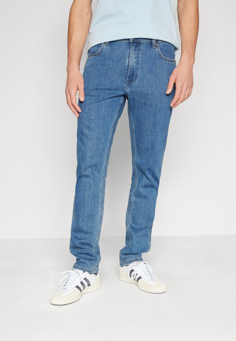 Jeans de mezclilla azul con un corte slim, diseño de cinco bolsillos, costuras visibles y un ligero desgaste. Combinados con zapatillas blancas con detalles en negro.
