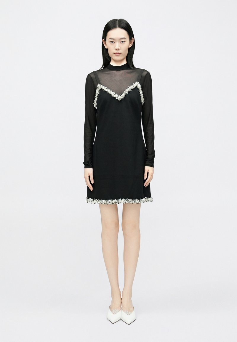 Melna kleita ar caurspīdīgu garām piedurknēm, rotāta ar ziedu akcentiem pie izgriezuma un apakšmalas, ar taisnu griezumu un gludu tekstūru.