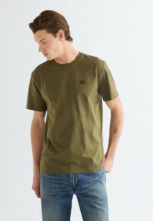 Giovane uomo con capelli castani corti, che indossa una maglietta verde oliva e jeans blu, guarda verso il basso sul lato sinistro.