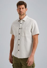 Camisa de botones de manga corta en beige claro con un patrón texturizado, que cuenta con dos bolsillos en el pecho y botones negros. Tejido duradero.