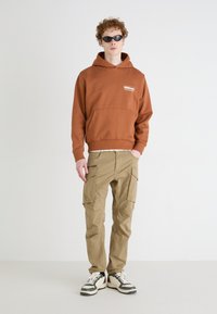 Bruine hoodie met logo, kangoeroezak, beige cargobroek met zijzakken, en zwart-witte sneakers. Model draagt zwarte zonnebril.