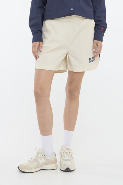 Beige shorts med elastisk linning, med en blå logo på venstre ben. Kombinert med kremfargede joggesko og hvite ankelsokker.