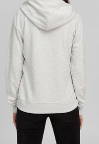 Persona che indossa una felpa con cappuccio grigia chiara e pantaloni neri, vista di spalle su uno sfondo neutro.