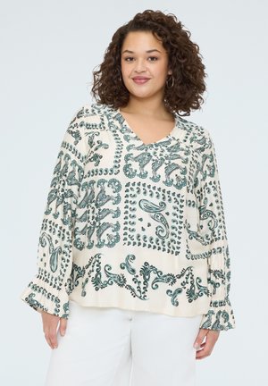 Lockige Frau trägt eine cremefarbene Bluse mit langen Ärmeln und dunkelgrünen Paisley-Mustern sowie weiße Hosen, lächelt leicht.