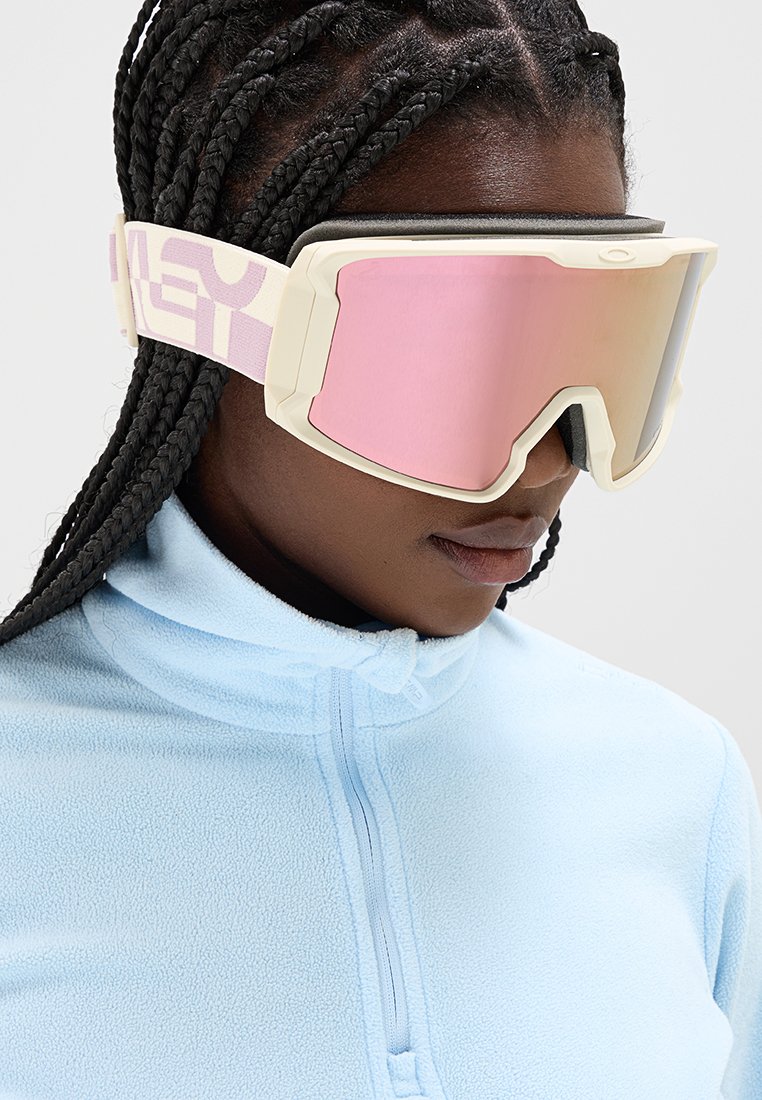 Skibrille mit einem weißen Rahmen und pinken, verspiegelten Gläsern. Der Riemen weist ein gemustertes Design auf und ist um einen hellblauen Fleece-Pullover befestigt.