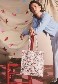 Moteris šviesiai mėlynu kardiganu, atsirėmusi į raudoną kėdę su gėlėtu pirkinių krepšiu, fone smėlio spalvos audinys su žvaigždžių raštu ir rožinis languotas grindų paviršius.