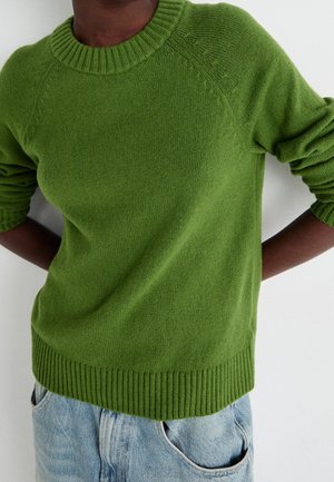 Osoba ubrana w zielony sweter z dzianiny z prążkowanymi mankietami i ściągaczem, w połączeniu z jasnoniebieskimi dżinsami, stojąca na jednolitym białym tle.