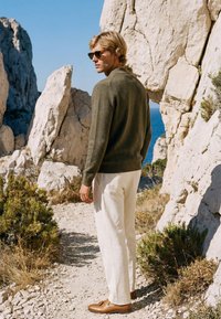Homme portant des lunettes de soleil, un pull vert, un pantalon blanc et des chaussures marron, debout sur un chemin rocheux près de grandes falaises blanches au bord de la mer sous un ciel dégagé.