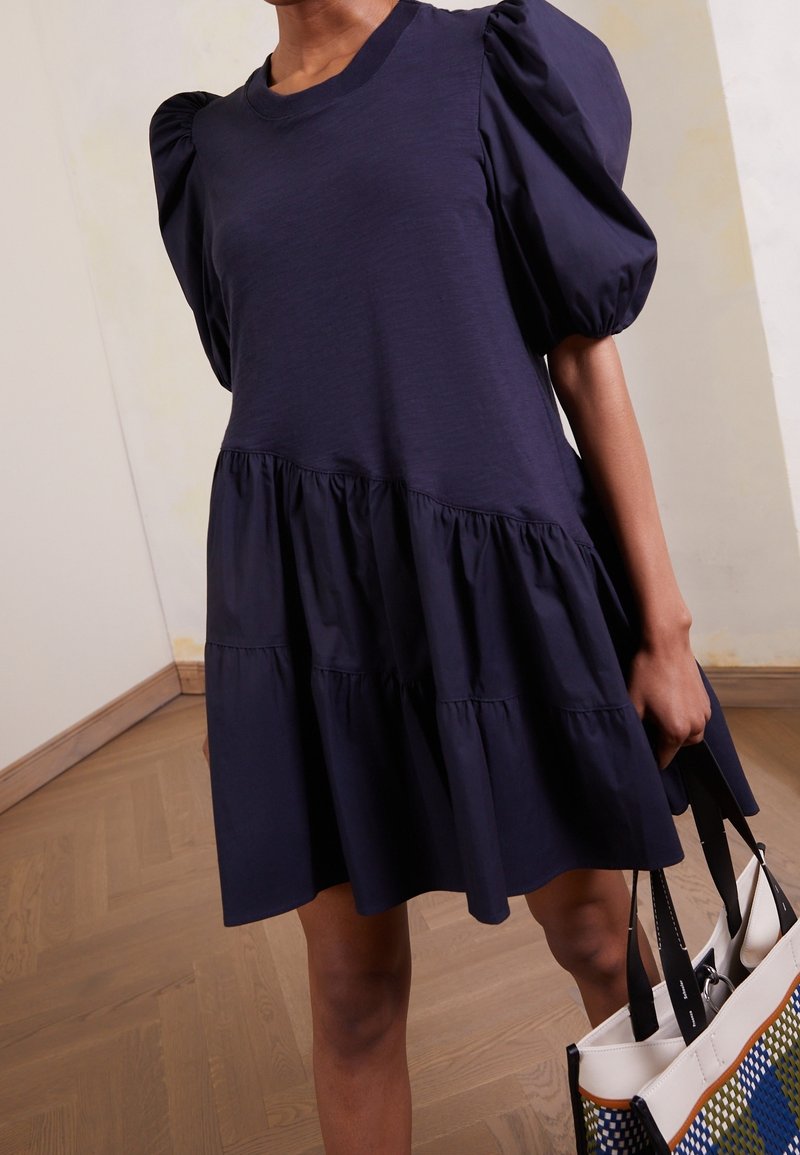 Navy blue dress with puff sleeves, tiered ruffles at the hem, made of soft fabric. Paired with a multicolored tote bag.