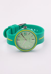 Reloj de silicona verde con una esfera redonda, acentos amarillos y marcadores de horas plateados. Tiene una correa texturizada y un cierre de metal.