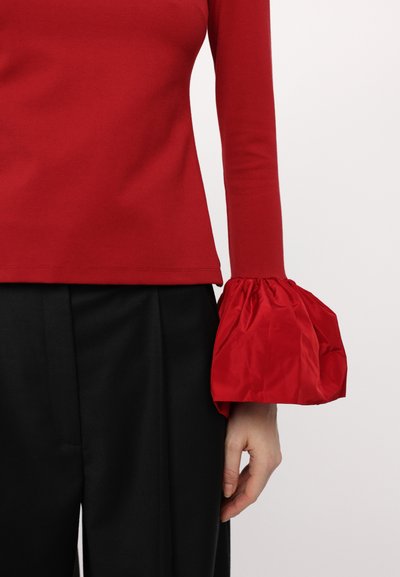 Red cotton top with long sleeves, featuring a voluminous red satin cuff. Paired with black trousers, highlighting contrasting textures.