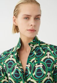 Bunt gemusterte Bluse mit einem auffälligen grünen und marineblauen Kreisdesign, aus glattem Stoff mit klassischem Kragen und Knopfdetails.