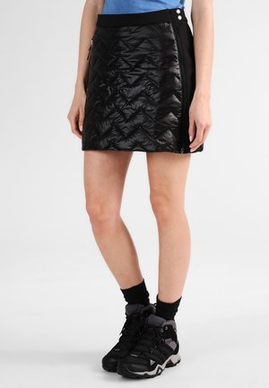 Femme portant une mini-jupe matelassée noire avec des boutons latéraux, des chaussettes noires, des chaussures de randonnée et un haut bleu, debout devant un fond uni.