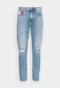 Jeans in denim azzurro chiaro con vestibilità slim, caratterizzati da toppe strappate, chiusura con bottone e un piccolo dettaglio di tasca rossa e bianca.