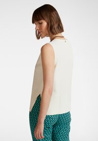 Top senza maniche di colore crema con pannelli laterali a coste, caratterizzato da un dettaglio sul retro e una vestibilità rilassata, abbinato a pantaloni verdi con fantasia.