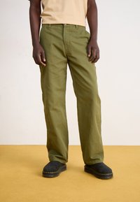 Olíva zöld pamut nadrág egyenes szabással, gombos záródással és oldalsó zsebekkel. Fekete velúr slip-on cipővel viselve.