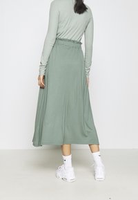 Pitkä, vaaleanvihreä mekko, jossa on pitkät hihat, kerätty vyötäröyksityiskohta ja virtaava hame, yhdistettynä valkoisiin urheilukenkiin ja sukkiin.