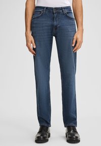 Niebieskie dżinsy o prostym kroju, z pięcioma kieszeniami i kontrastowym przeszyciem. Noszone z czarnymi butami, charakteryzującymi się gładką teksturą i trwałym materiałem.