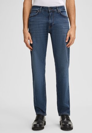 Niebieskie dżinsy o prostym kroju, z pięcioma kieszeniami i kontrastowym przeszyciem. Noszone z czarnymi butami, charakteryzującymi się gładką teksturą i trwałym materiałem.