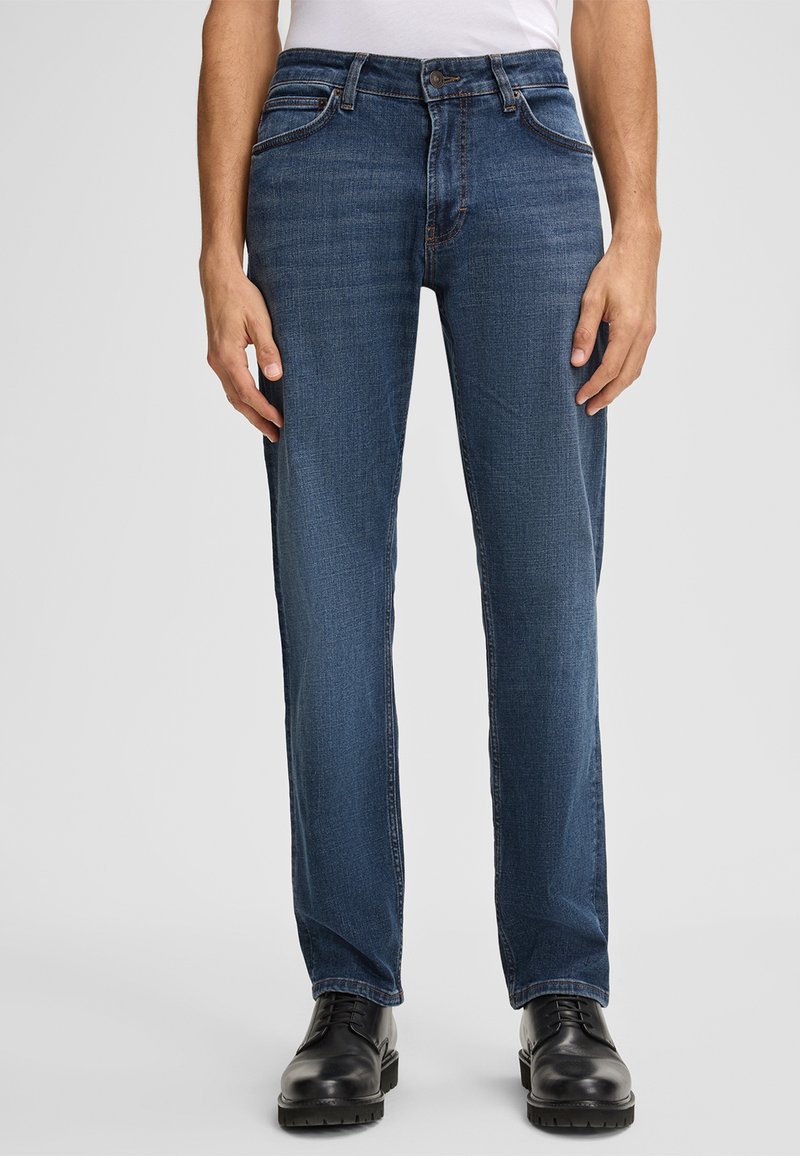 Niebieskie dżinsy o prostym kroju, z pięcioma kieszeniami i kontrastowym przeszyciem. Noszone z czarnymi butami, charakteryzującymi się gładką teksturą i trwałym materiałem.