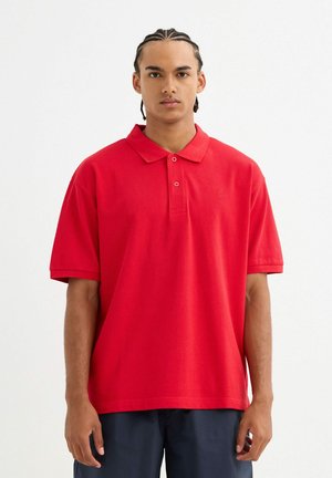 Camisa polo de algodón roja con cuello, mangas cortas y botonera de dos botones; presenta un sutil logo en el lado izquierdo del pecho.