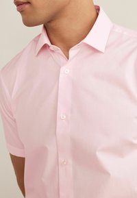 Gros plan sur un homme portant une chemise rose clair à manches courtes, boutonnée et avec un col, visible des épaules au menton.