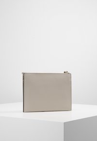 Pochette rectangulaire en cuir beige à surface texturée, avec tirette de fermeture éclair dorée, posée sur un socle cubique blanc sur un fond uni.