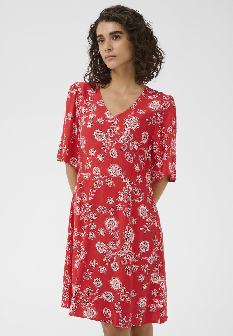 Femme aux cheveux bouclés et foncés portant une robe rouge à manches courtes avec un motif floral blanc, debout devant un fond blanc uni.
