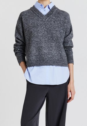 Personne portant un pull en tricot gris à col en V sur une chemise bleu clair à col, associée à un pantalon noir, debout avec une main dans la poche.