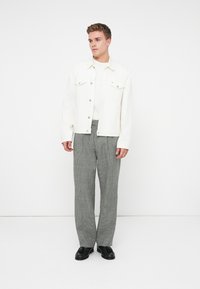 Fiatal férfi fehér farmerdzsekit, fehér inget, szürke kockás nadrágot és fekete bőr loafert visel, fehér háttér előtt állva.