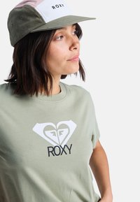 Zelené bavlněné tričko s bílým srdíčkovým logem a nápisem "ROXY" v černé barvě. Doplňuje ho dvoubarevná kšiltovka s růžovými, zelenými a bílými panely.