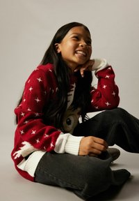 Jeune fille souriante aux longs cheveux noirs assise par terre, portant un pull rouge et blanc à motifs et un pantalon foncé, sur un fond uni.