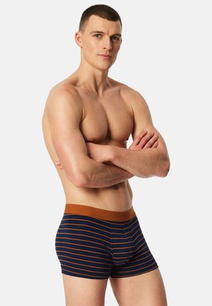 Shirtless man with short hair, arms crossed, wearing navy blue boxer briefs with horizontal orange stripes and an orange waistband.