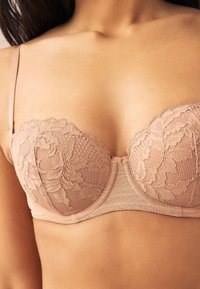 Primo piano di una donna che indossa un reggiseno con ferretto in pizzo color nude con motivi floreali e spalline regolabili.