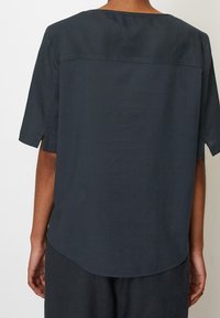 Tmavě šedá košile s krátkým rukávem a volným střihem, vyrobená z lehké látky; má kulatý výstřih a mírně zakřivený zadní lem.