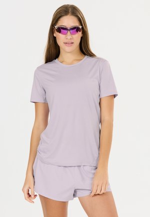 Femme portant un t-shirt de sport violet clair et un short, avec des lunettes de sport violettes réfléchissantes, posant devant un fond blanc uni.