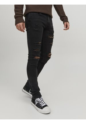 Homme portant un jean skinny noir déchiré, des baskets montantes noires avec des lacets blancs et un pull en tricot marron, debout devant un fond uni.