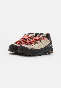 Chaussures de randonnée beiges et noires avec des lacets orange, tige en daim texturé, semelle en caoutchouc robuste et embout renforcé. Marque Vibram visible.