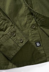 Camisa de botones color verde oliva con una textura suave, que cuenta con dos botones en el puño y una etiqueta negra con un logo blanco en el lateral.