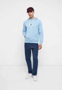 Γαλάζια φουτεράκη με λευκό λογότυπο, μπροστινές τσέπες και κορδόνια σύσφιξης. Συνδυάζεται με σκούρο μπλε τζιν και λευκά αθλητικά παπούτσια. Φοράει καπέλο μπορντώ.