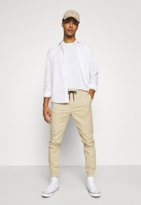Beige jogger pants with elastic cuffs, paired with a white t-shirt and overshirt. White high-top sneakers and a light-colored cap complete the look.