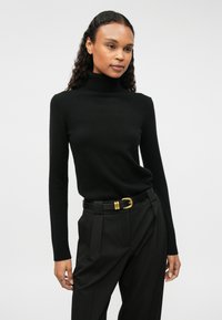 Μαύρο πουλόβερ με ψηλό λαιμό σε μαλακή υφή, συνδυασμένο με μαύρα παντελόνια ψηλόμεσα και μαύρη ζώνη με χρυσό κούμπωμα.