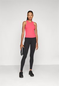 Top sportivo rosa con colletto alto, abbinato a leggins neri. Il modello tiene in mano una borraccia nera e indossa scarpe da ginnastica nere.