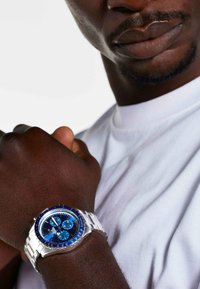 Silver metal wristwatch with a blue dial, white and blue accents, three subdials, and a stainless steel bracelet, worn on a dark-skinned wrist.