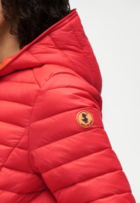 Γυναίκα που φοράει κόκκινο καπιτονέ μπουφάν με κουκούλα, με στρογγυλό πορτοκαλί ετικέτα λογότυπου «Save the Duck» στο πάνω μέρος του μανικιού.